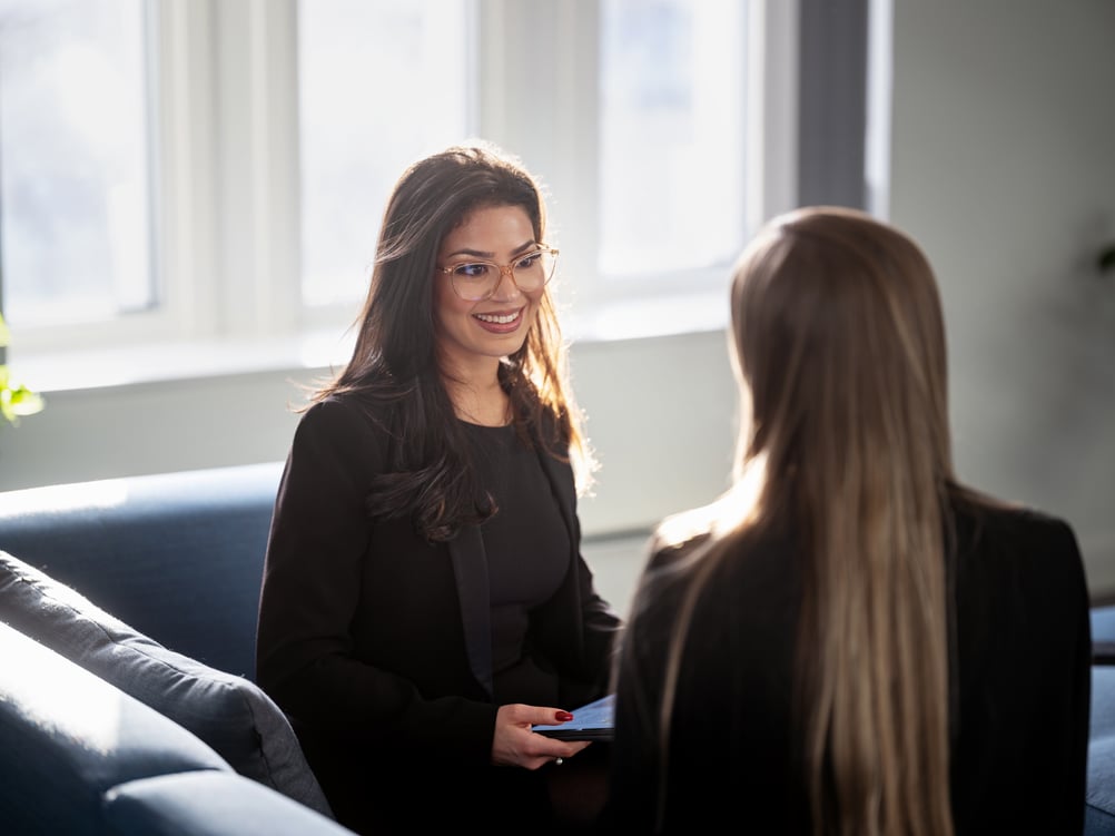 Two colleagues chatting while sitting on a couch in a modern office setting.