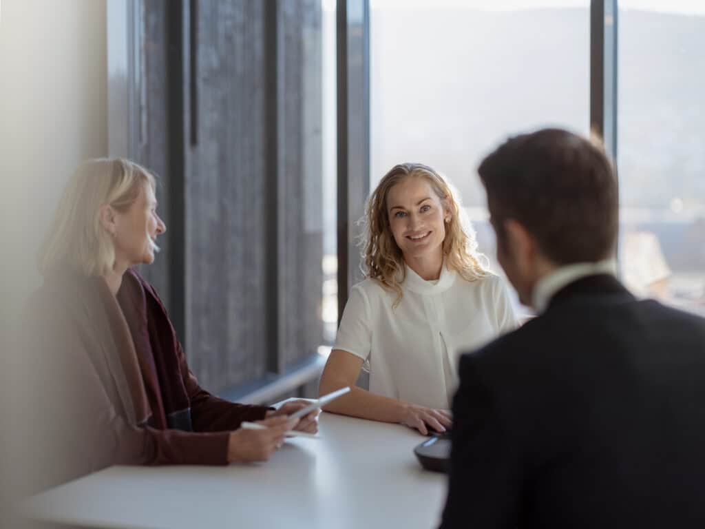 Three colleagues are seated at a table in a professional office setting, engaged in discussion.