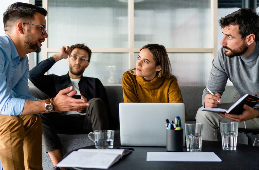 Fire personer i gang med en diskussion omkring et bord, med en åben bærbar computer foran dem.