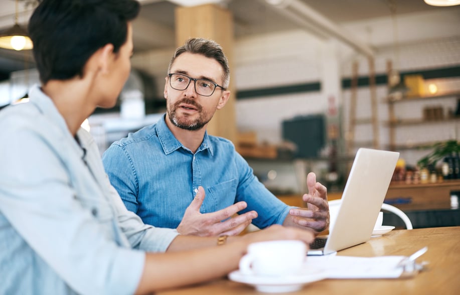 A man and woman are at a table, using a laptop, appearing to collaborate or engage in a discussion.