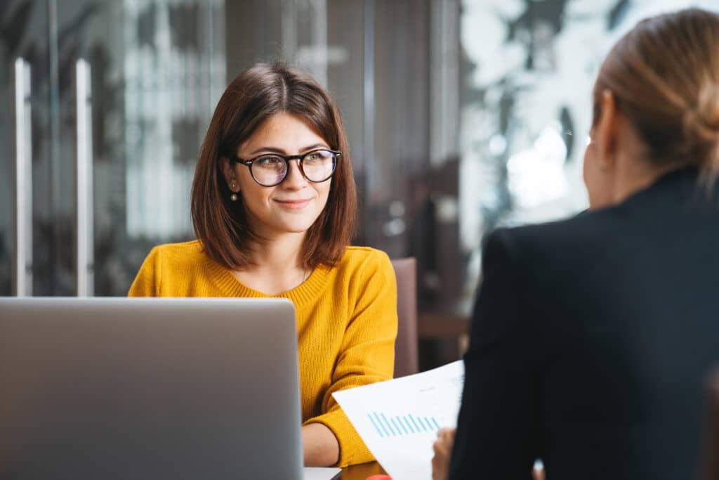 Girl in yellow jumper and glasses in a meeting room with another colleague.