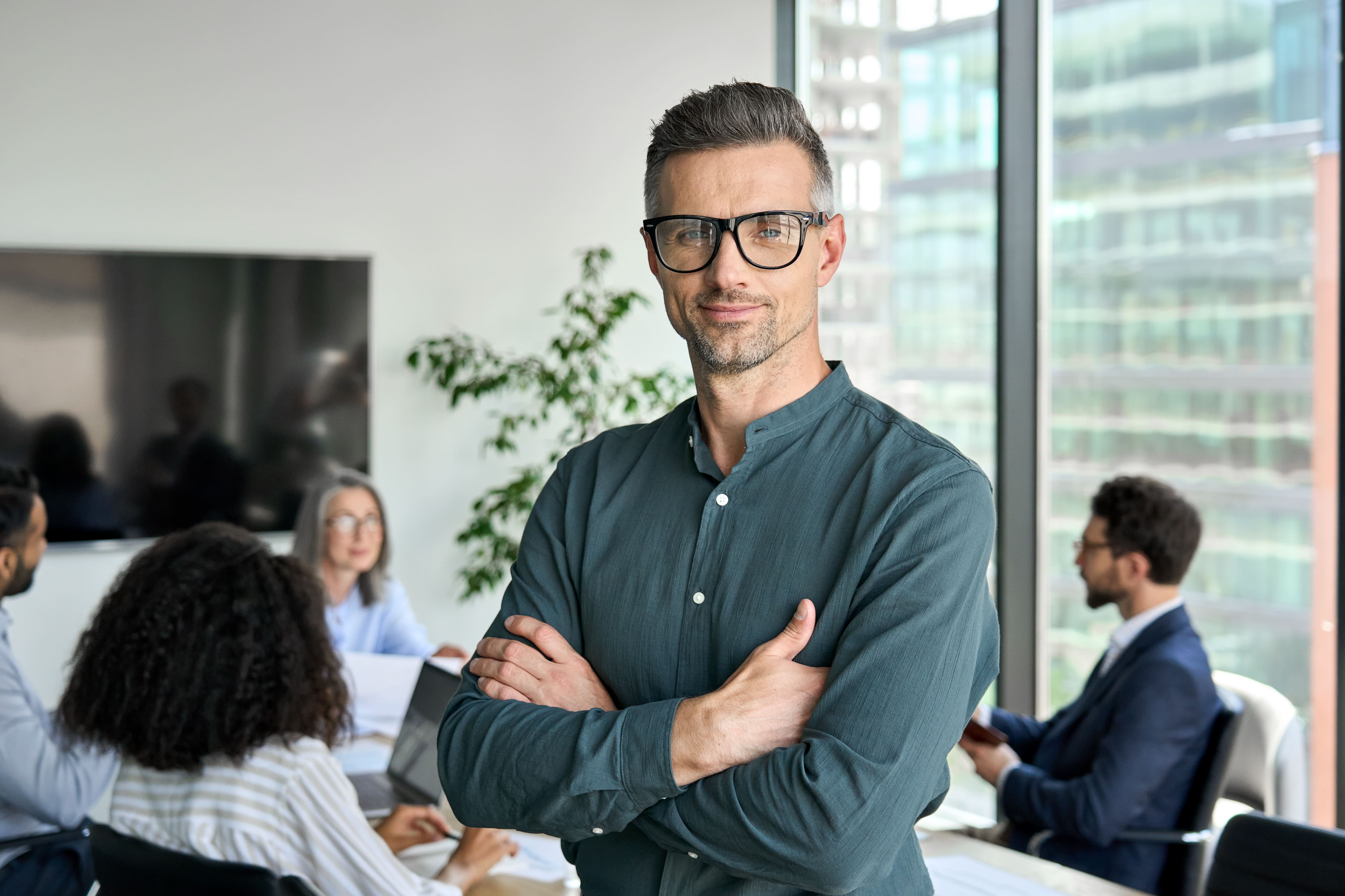 A man wearing glasses stands in front of a group of people in a meeting room, engaged in discussion.