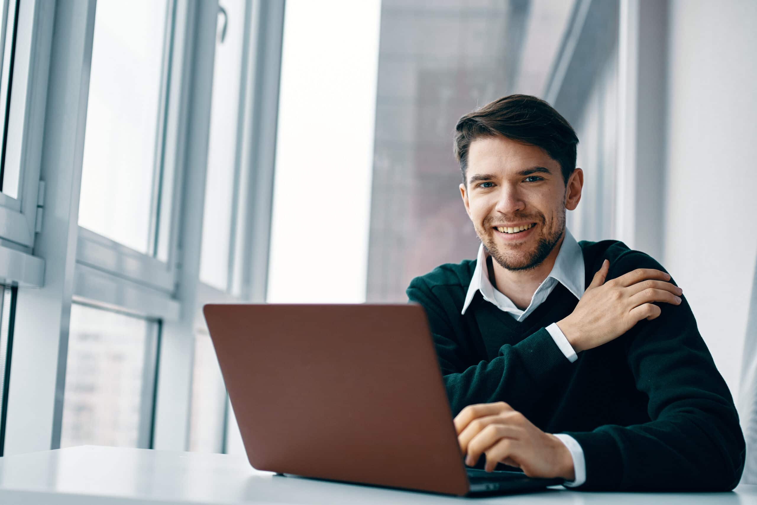 Smiling man in dark sweater and white collar shirt working on laptop near large office windows with natural light.