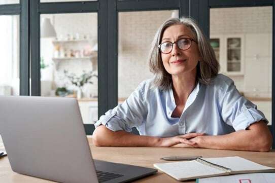 En kvinna med glasögon sitter vid ett skrivbord och är engagerad med sin laptop