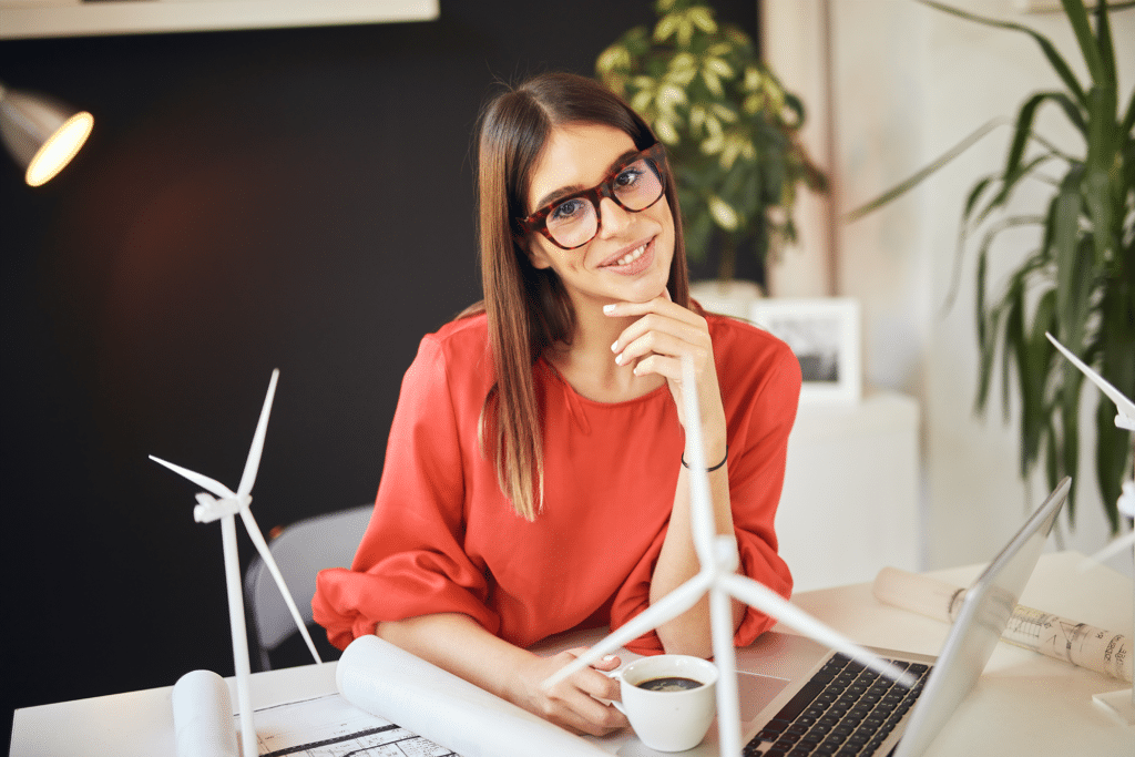 A woman sits at a desk, holding a coffee cup, with her laptop in front of her.