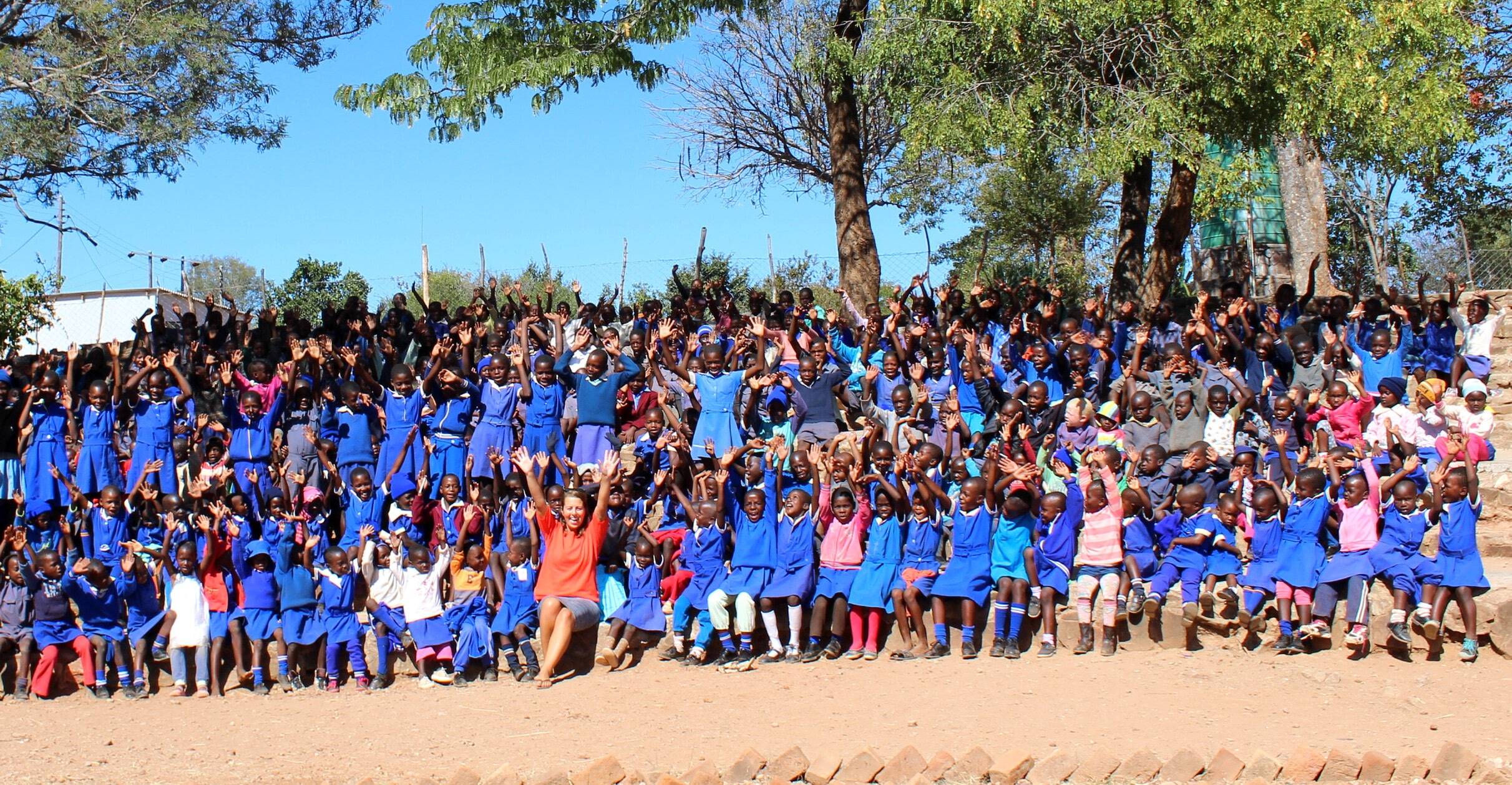 A large group of Zimbabwean school children in blue uniforms wave and smile at the camera