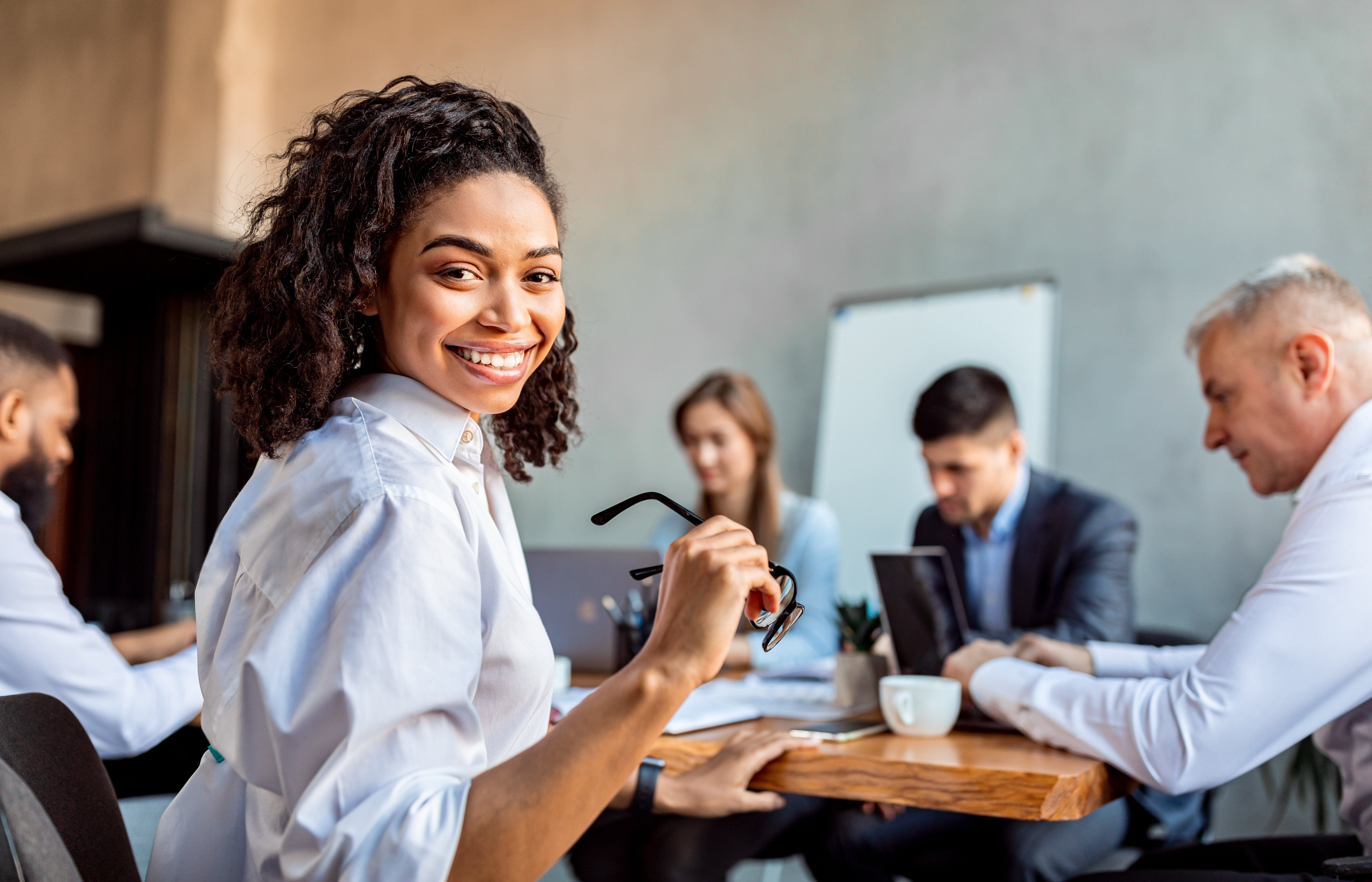 A smiling woman sits at a table with colleagues in a meeting room, engaged in a discussion