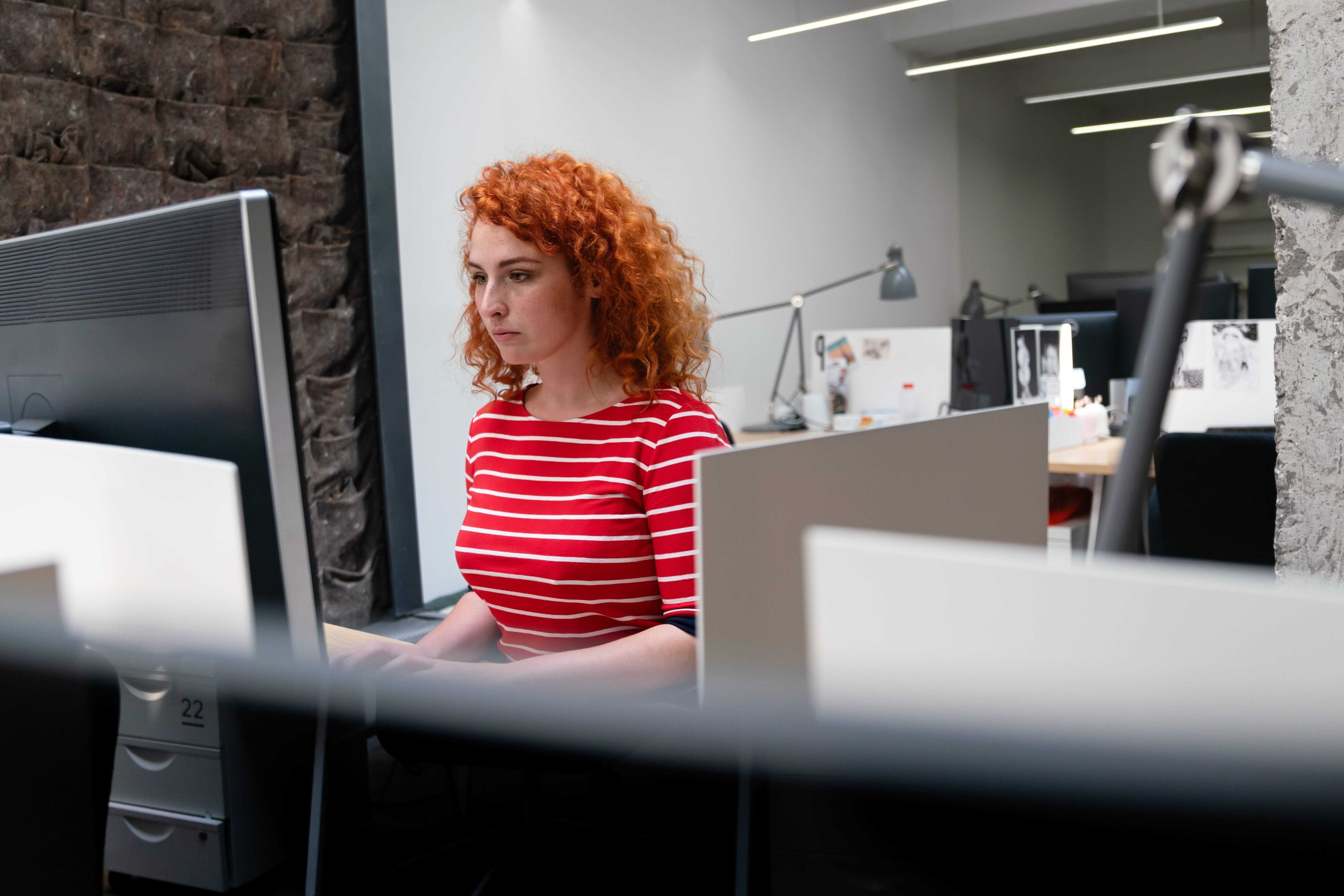 A woman is working at her desk, focusing on a large computer monitor in an open office space